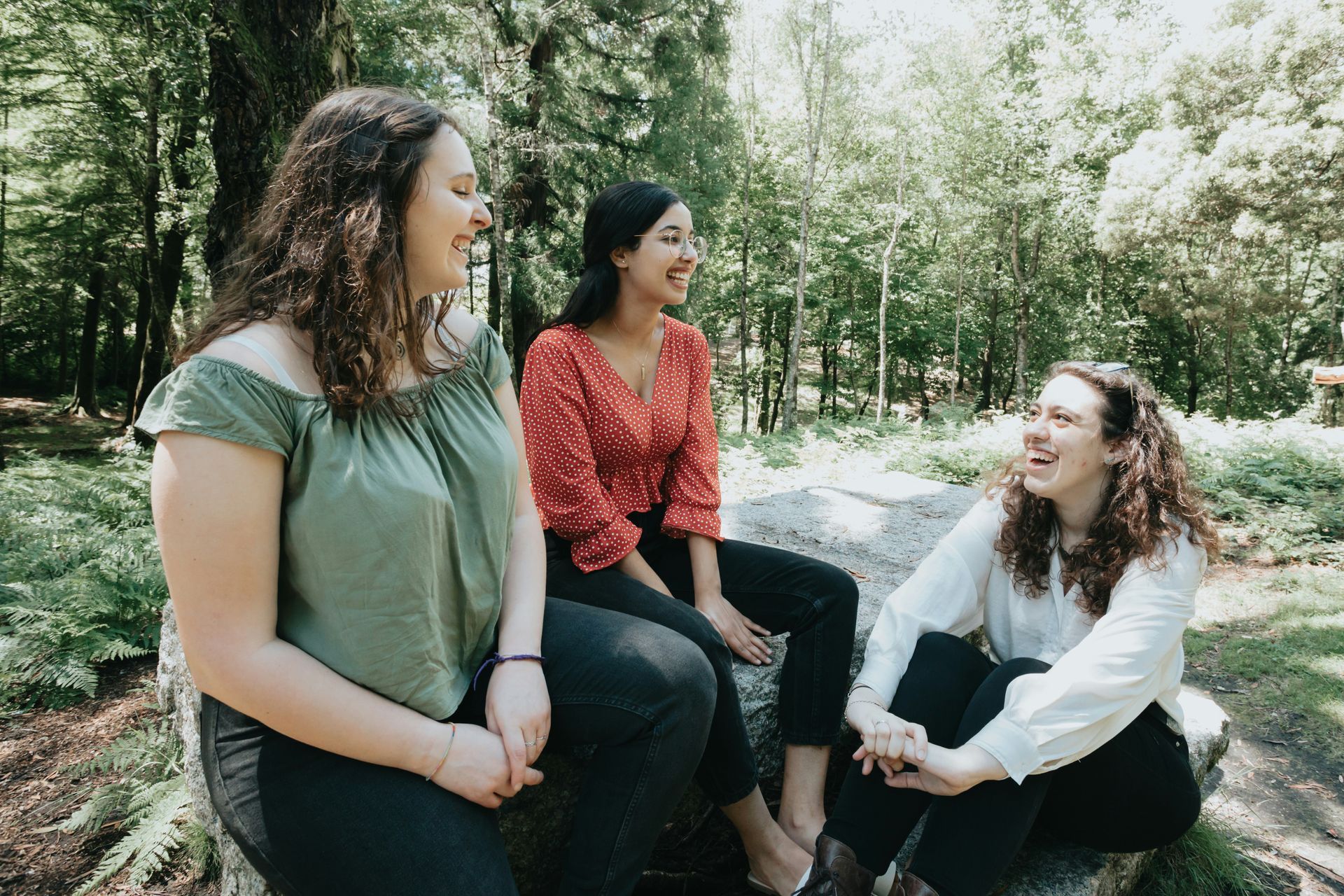 Three people laughing while sitting on a rock in a wooded area.