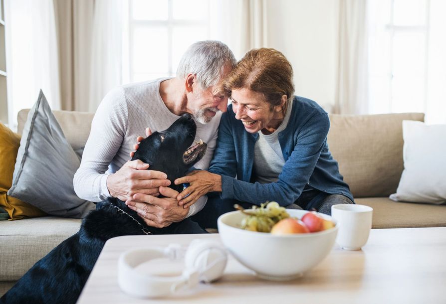 Senior couple on sofa petting a black dog, laughing, with fruit bowl on coffee table.