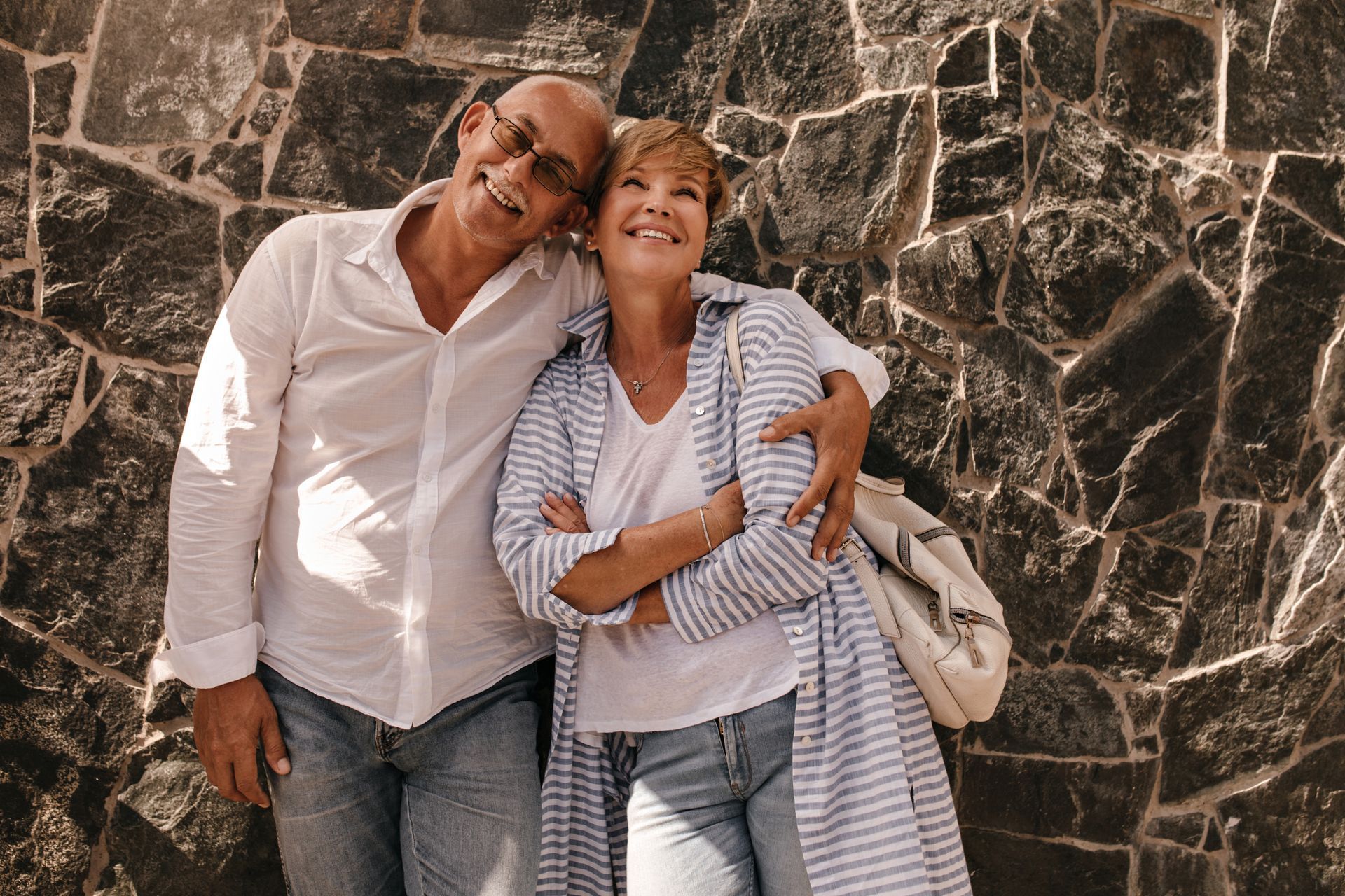 Man with arm around woman, both smiling against stone wall.