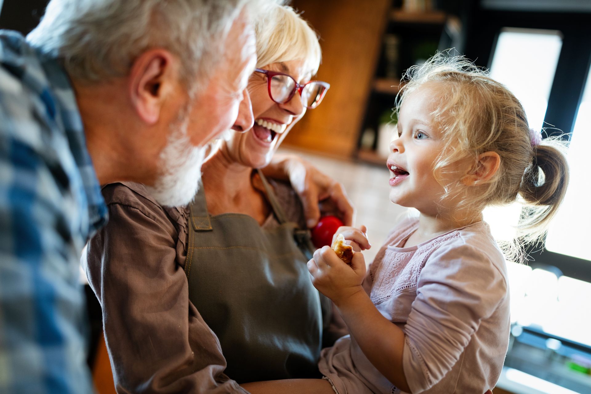 Elderly couple laughing with a young child in a kitchen, natural light.