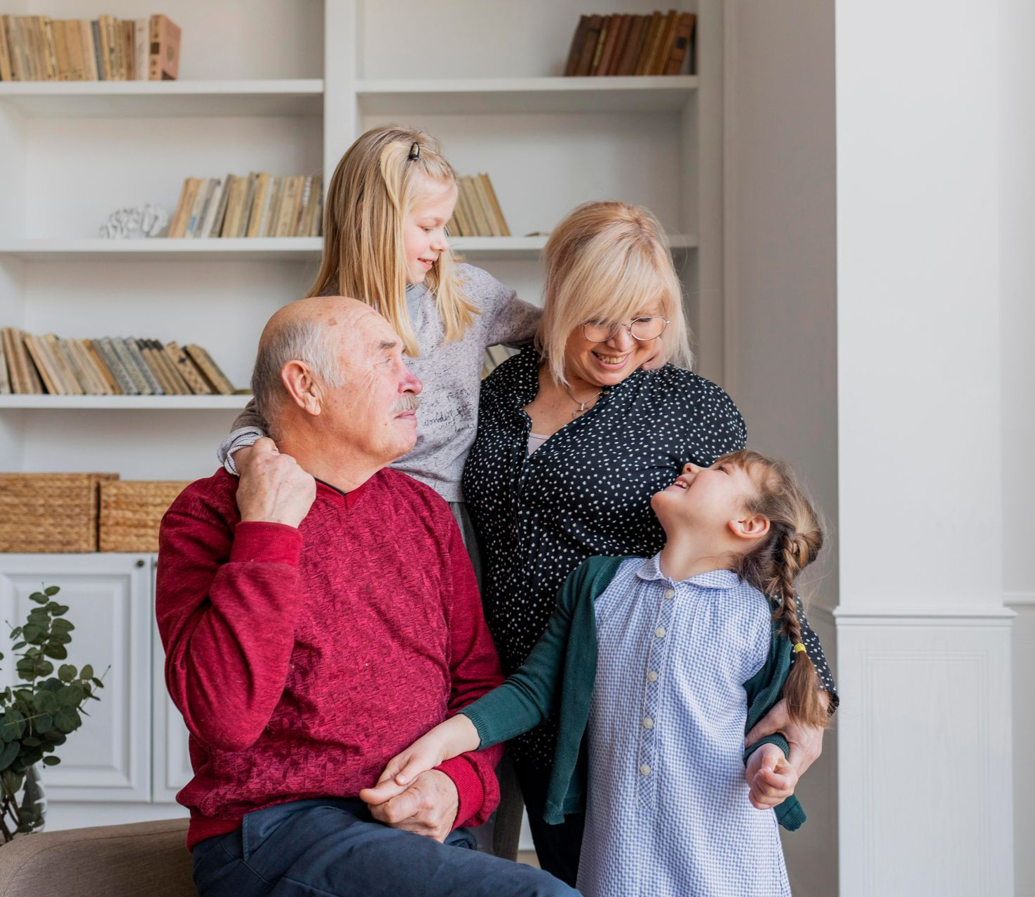 Grandparents with two granddaughters in a living room, smiling, arm around each other, bookshelf in background.