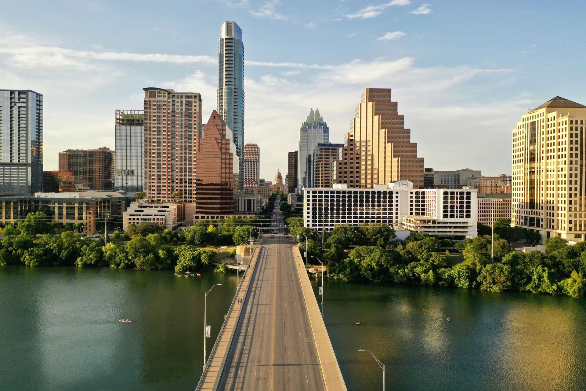 Austin, Texas skyline with bridge over green water, blue sky. Modern buildings and trees line the waterfront.