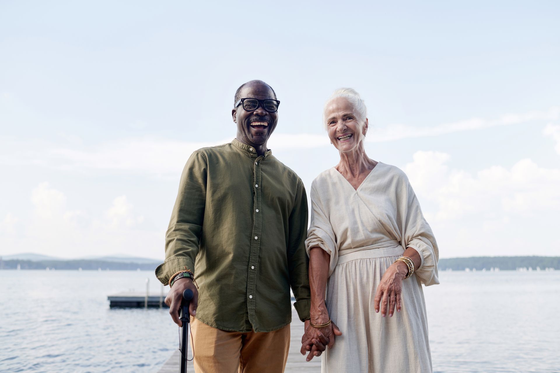 Couple holding hands, smiling. Man with cane, woman in white dress, standing near water.