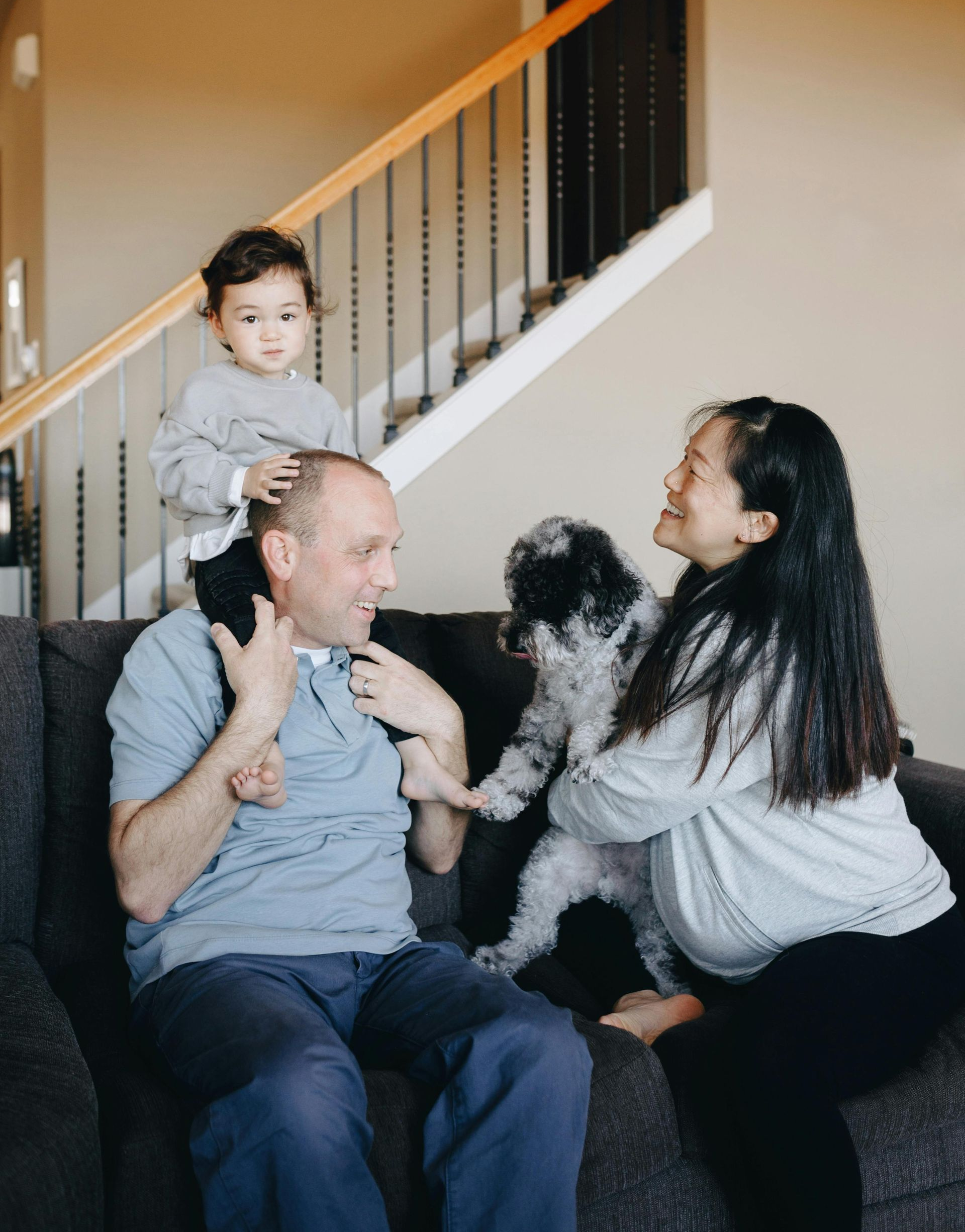 Family of four, including a small child on father's shoulders, and a dog, smiling on a sofa.