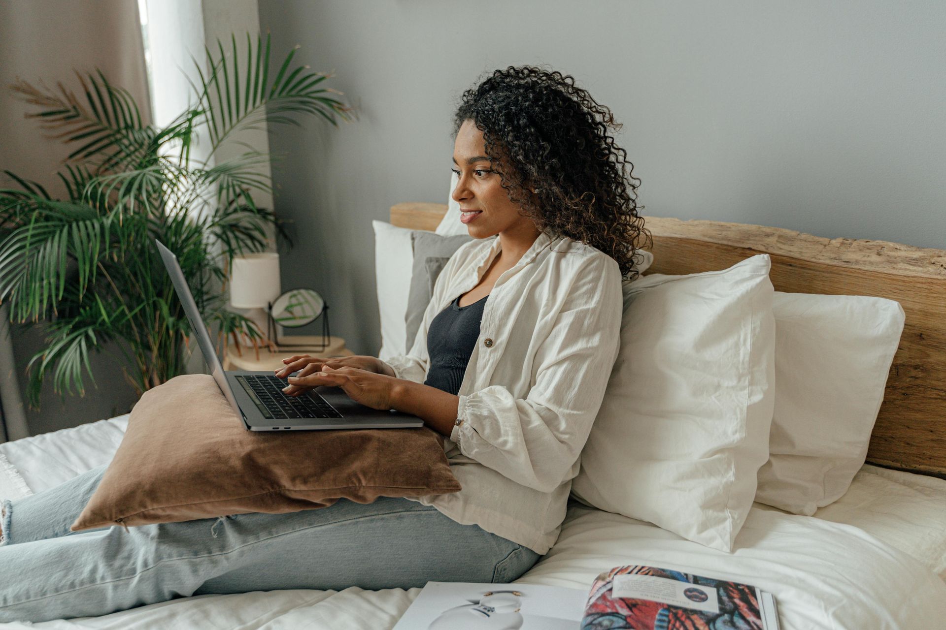 Woman using a laptop while sitting on a bed with pillows. There is a plant and side table in the background.