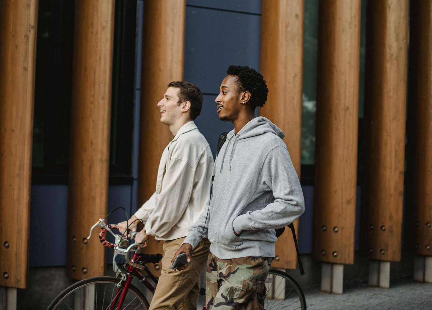 Two people walking near a wooden building, one holding a bicycle and smiling.