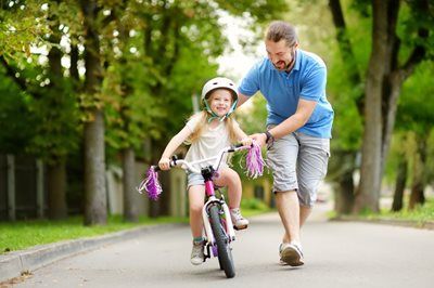A person runs alongside a child who is riding a bicycle on a paved path lined with trees.