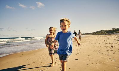 Two children run joyfully along a sandy beach toward the camera, with two people walking in the distance near the surf.