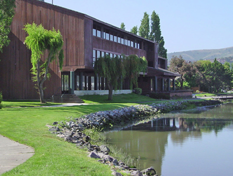 A multi-story brown wooden building sits on a green lawn beside a calm lake with a rocky shoreline.