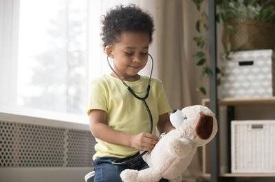 A child wearing a stethoscope plays doctor by listening to the chest of a stuffed dog toy in a bright, indoor setting.