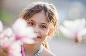A smiling child peeks through soft, out-of-focus magnolia flowers in a sunlit outdoor setting.