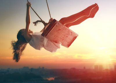 A person swinging on a wooden swing against a sunset, silhouetted above a cityscape.