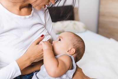A person in a white shirt sits on a white bed while breastfeeding an infant.