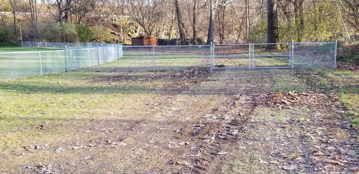 A chain-link fence surrounds a grassy area with fallen leaves, gate open, with trees in the background.