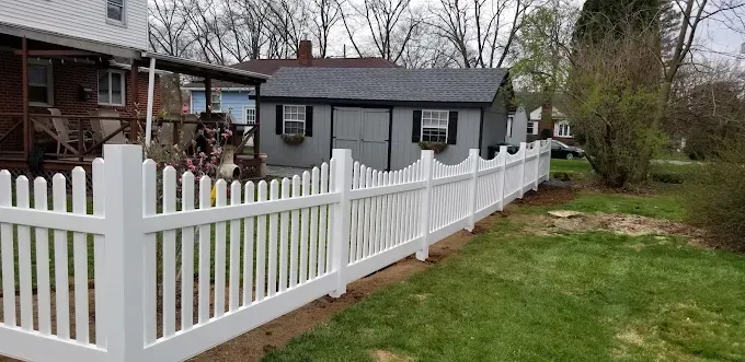 White picket fence in a yard next to a house and a shed. Overcast sky.