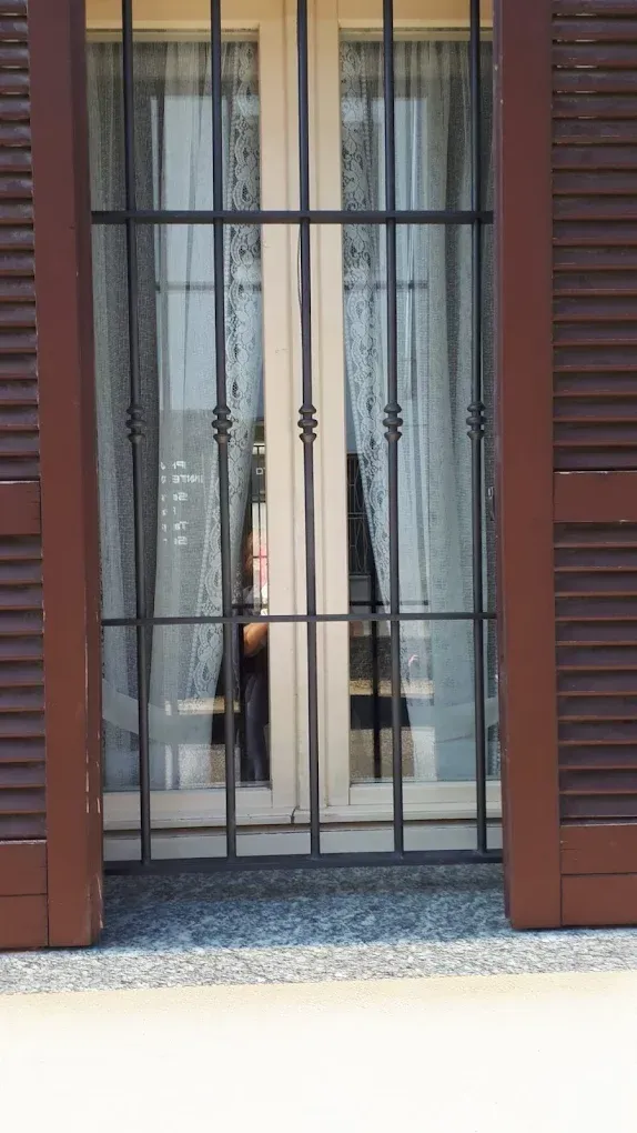 Finestra con sbarre di metallo scuro, telaio beige, persiane marroni e tende bianche trasparenti.