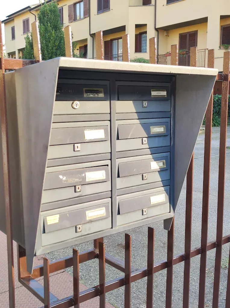 Gruppo di cassette postali grigie su un cancello marrone, all'aperto di fronte a un edificio.