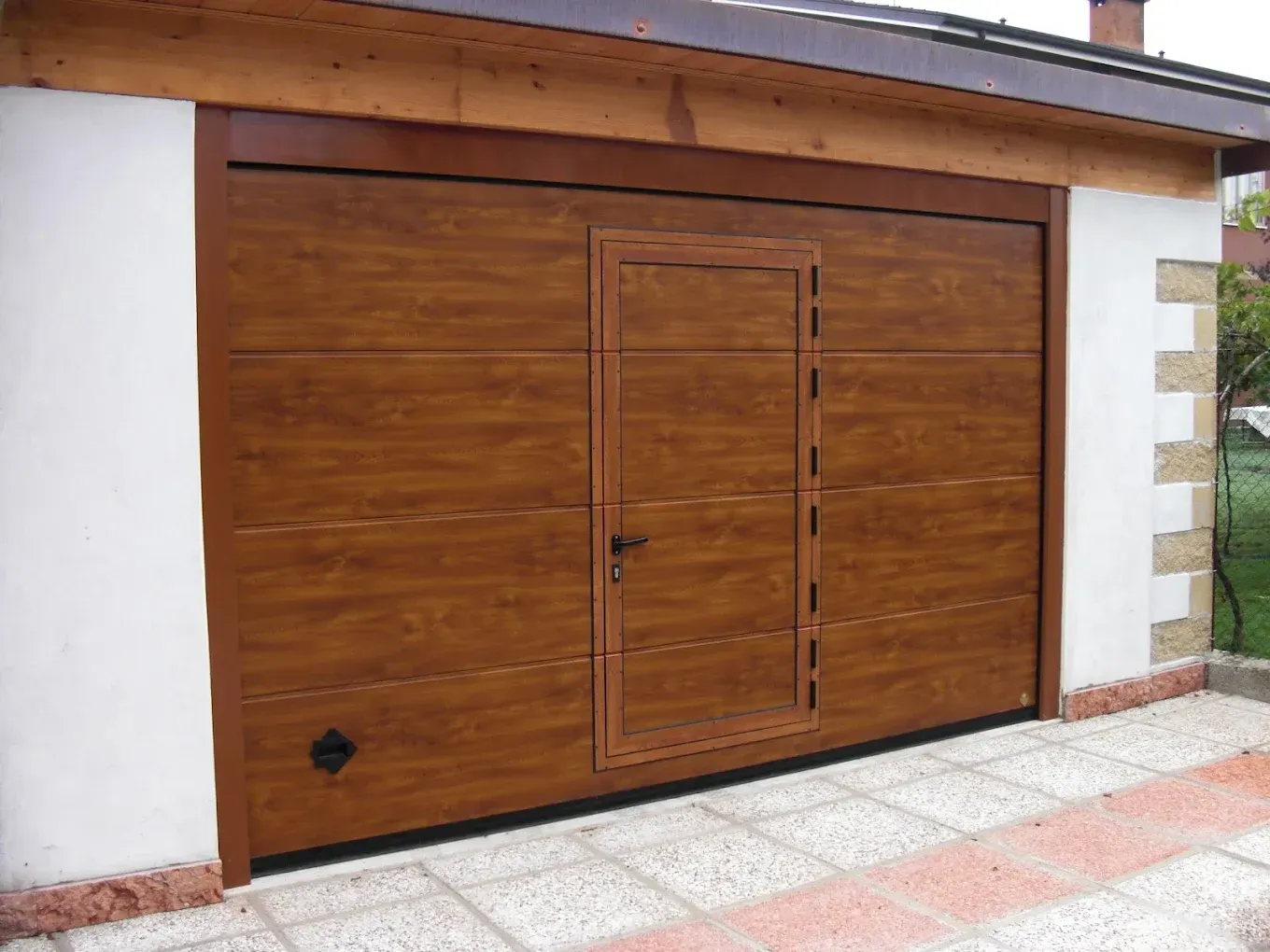 Portone da garage marrone con porta pedonale integrata. Esterno in mattoni bianchi e beige.
