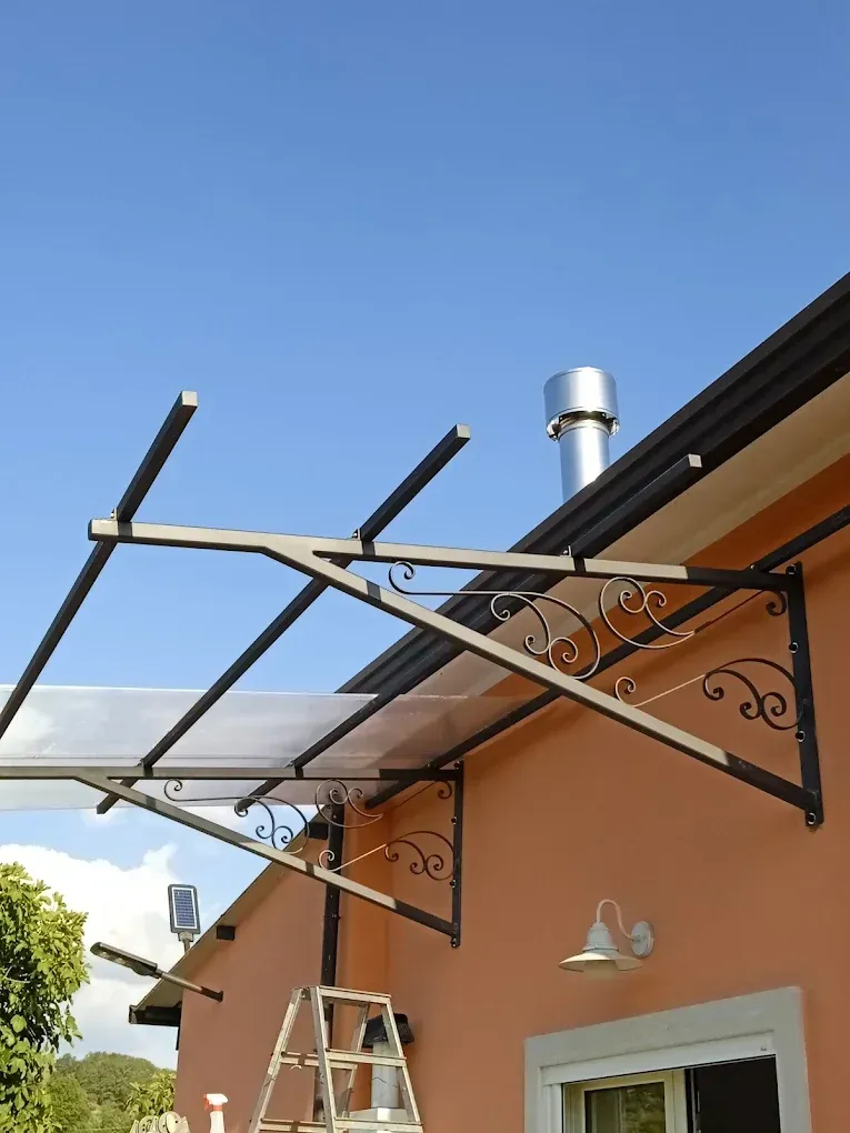 Tenda da sole in metallo con staffe decorative sopra una finestra e una porta. Sullo sfondo, un cielo azzurro brillante.