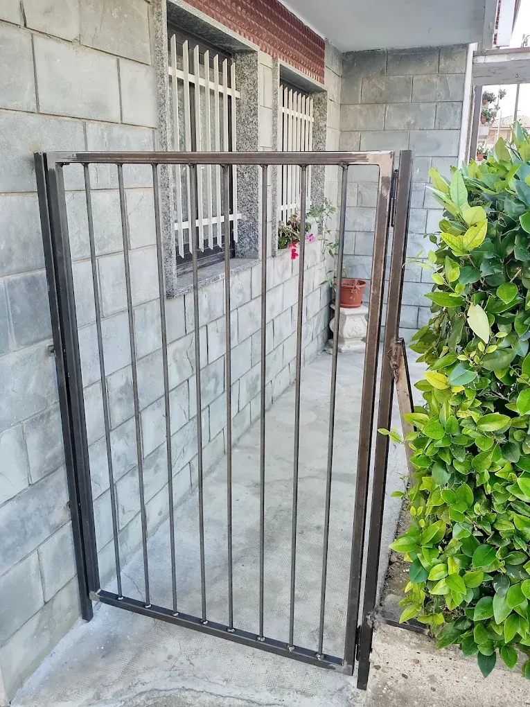 Cancello metallico, parzialmente aperto, in uno stretto spazio esterno. Sono visibili muri in blocchi grigi, finestre e vegetazione.