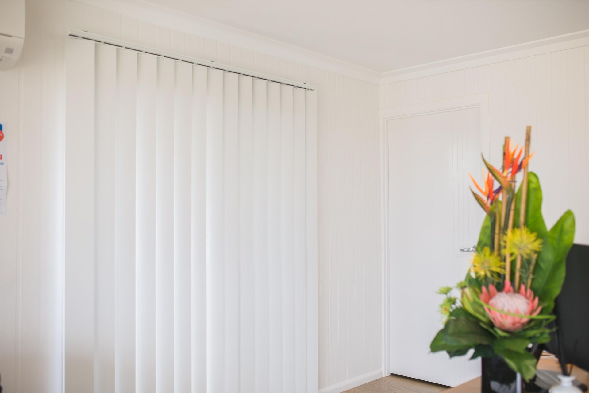 A Living Room With White Vertical Blinds And A Vase Of Flowers — Wares Blinds, Screens & Awnings In Gladstone Central, QLD