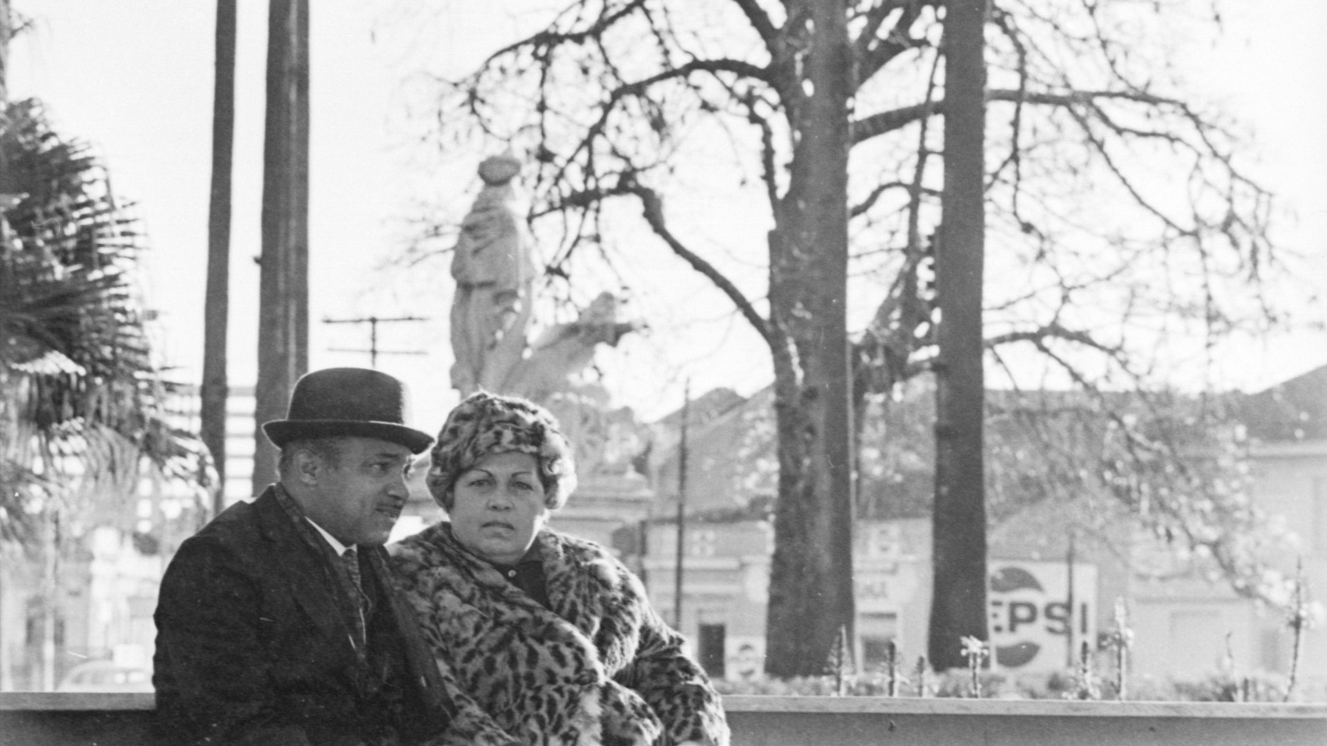 A man and woman are sitting on a bench in front of a pepsi sign.