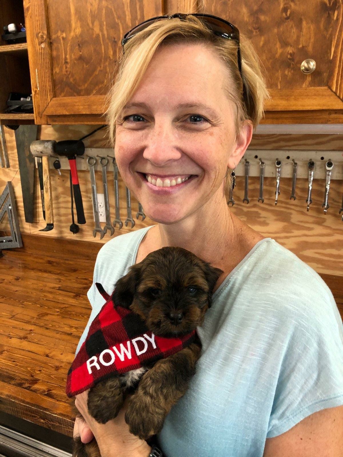 A woman is holding a puppy with a bandana that says rowdy on it.