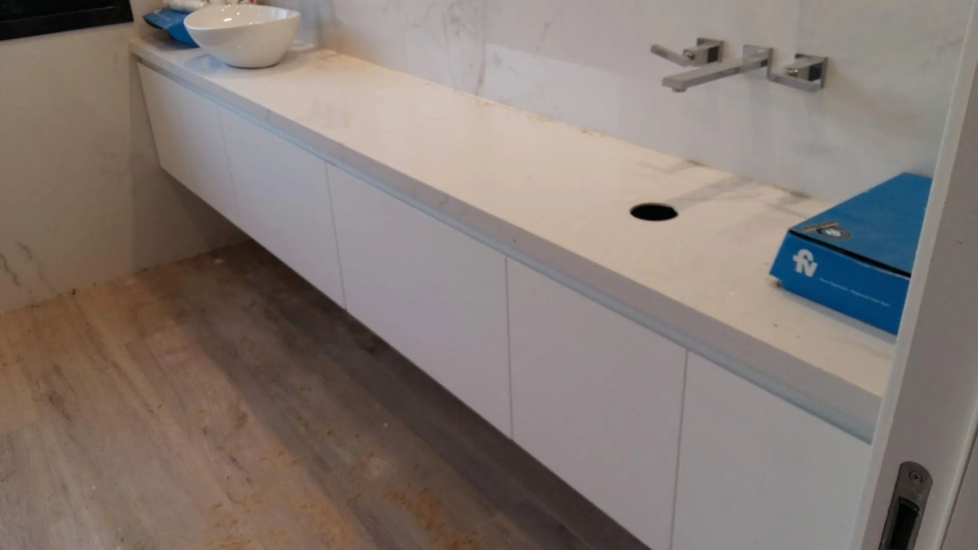 Baño moderno con una larga encimera blanca, lavabo, papelera azul y suelo de madera clara.