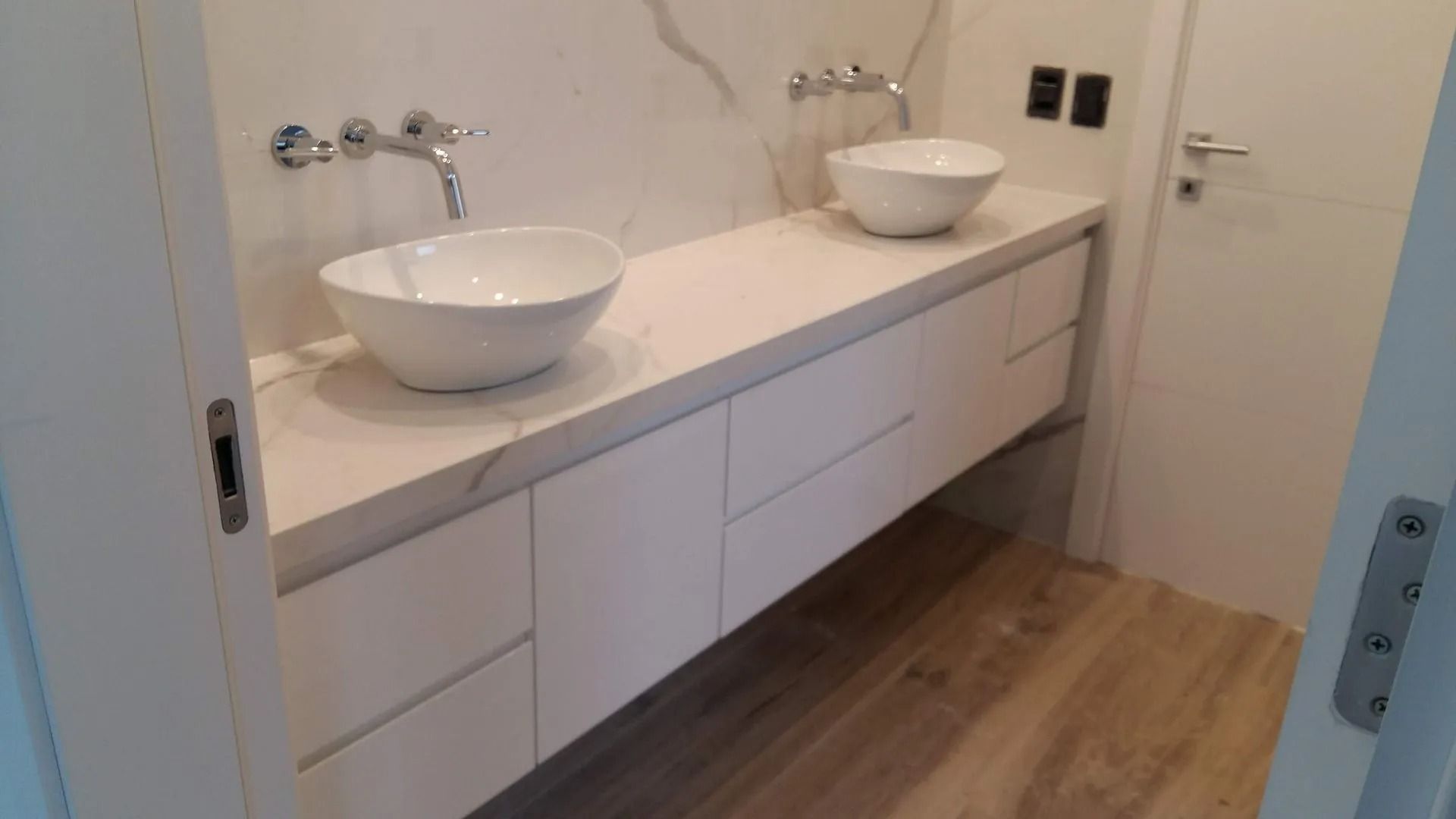 Tocador de baño moderno con dos lavabos blancos sobre encimera, armarios blancos y suelo de madera.
