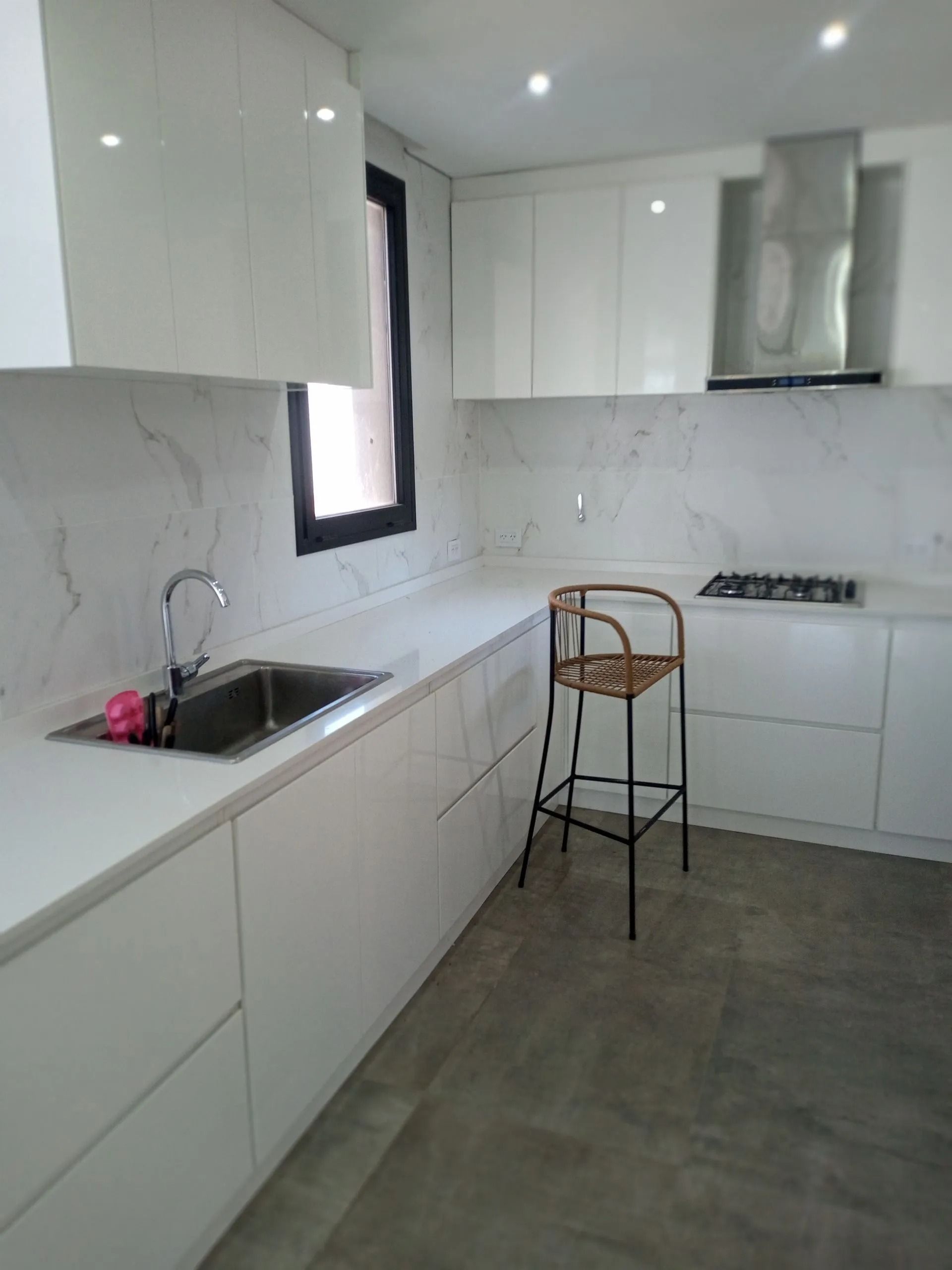 Cocina moderna blanca con detalles en negro, suelo gris, fregadero, vitrocerámica y una silla alta junto a la encimera.