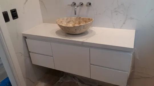 Tocador de baño moderno con cajones blancos, encimera de mármol y lavabo redondo de piedra.