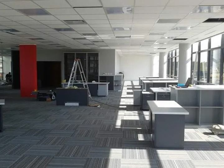 Espacio de oficina vacío con moqueta modular, escritorios, una columna roja y luz solar que entra por grandes ventanales.