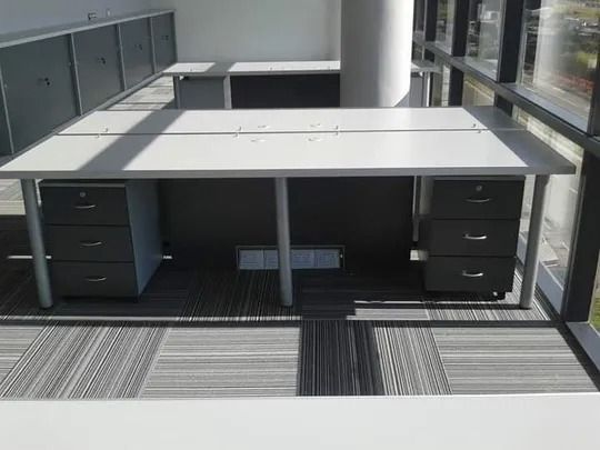 Cubículos de oficina modernos con escritorios grises, archivadores negros y alfombra a rayas cerca de grandes ventanales.