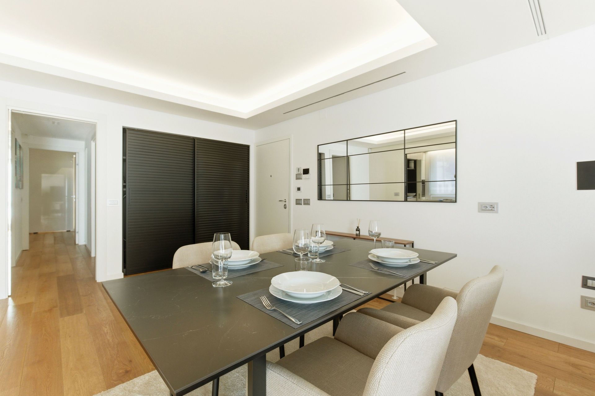Comedor moderno con mesa negra, sillas beige, espejo de pared y suelo de madera.
