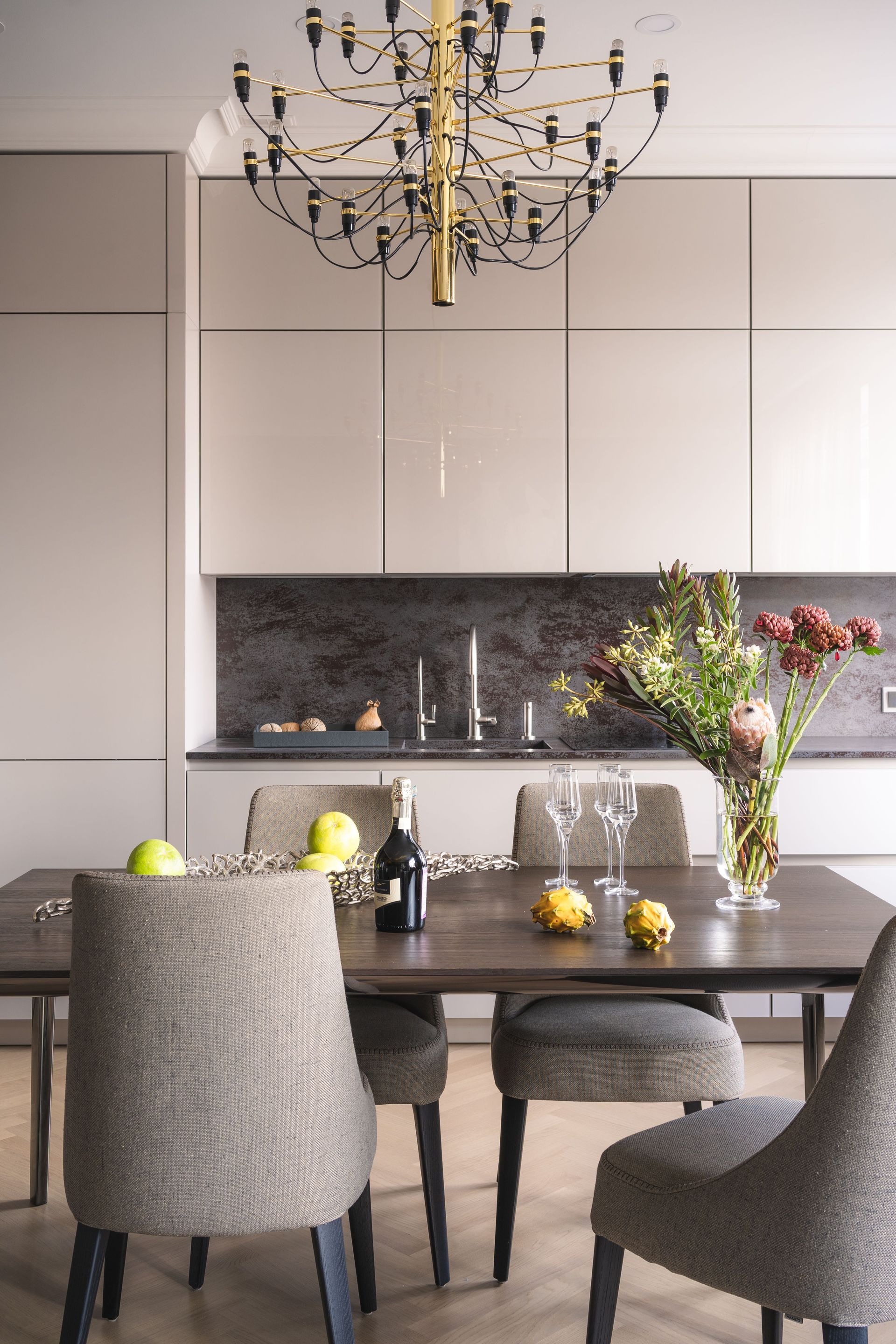 Comedor moderno con sillas grises, mesa oscura, armarios blancos, salpicadero negro y lámpara de araña.