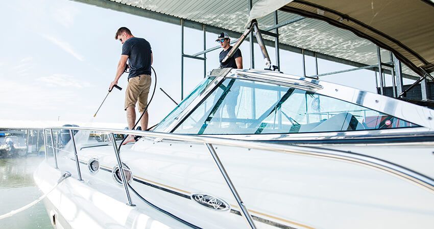 Two people washing a white boat with a pressure washer, under an awning on a sunny day.