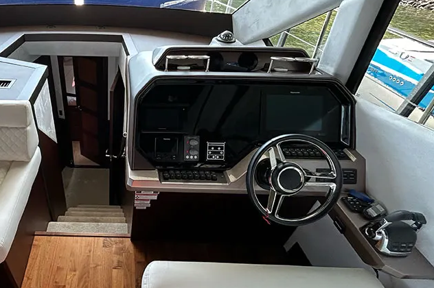 Boat's interior: helm with steering wheel, navigation screens, and controls. Dark colors, view of outside pier.