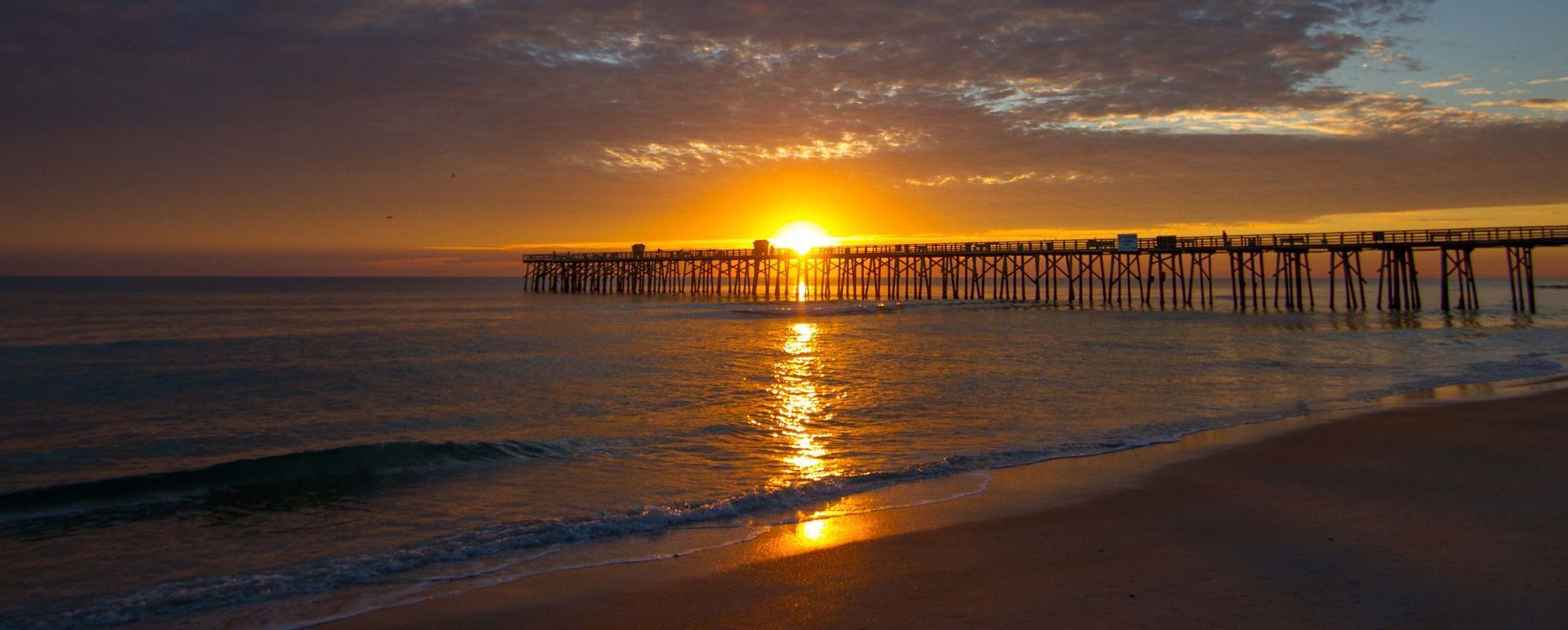 Sunset over ocean with a pier stretching out into the water, reflected in the waves and wet sand.