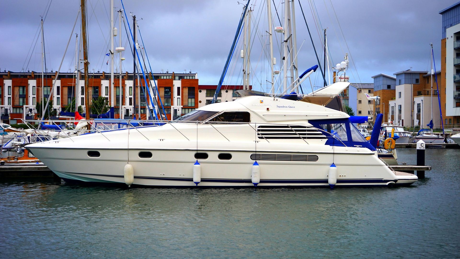 White motor yacht docked in a harbor, with blue accents and buildings in the background under a cloudy sky.