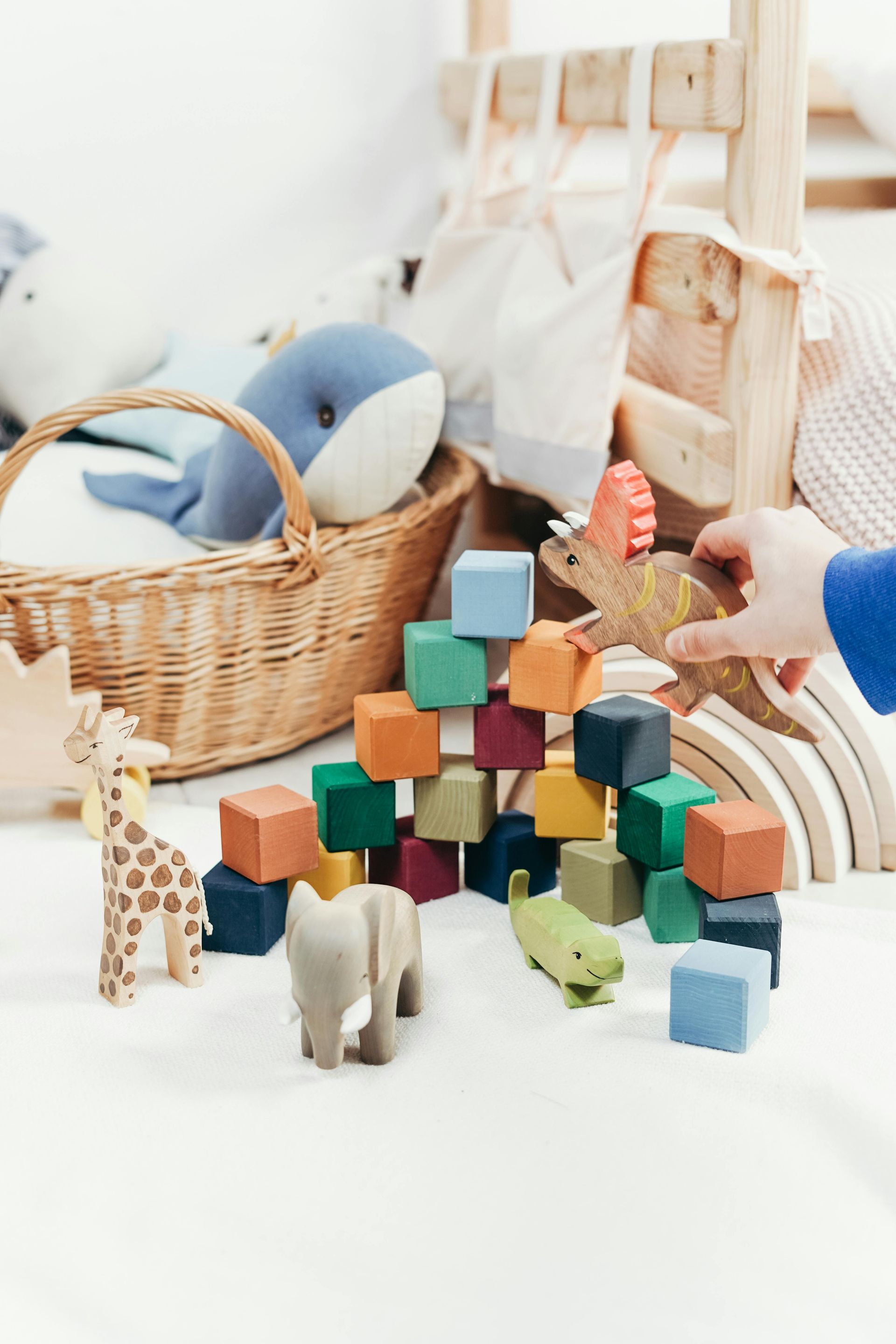 Assortment of wooden toys, including blocks, animals, and a rainbow arch on a white surface. A hand holds a dinosaur.