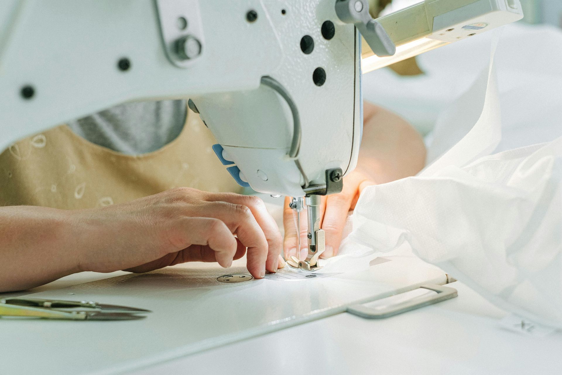 Hands guiding white fabric under a sewing machine needle; scissors on the table.