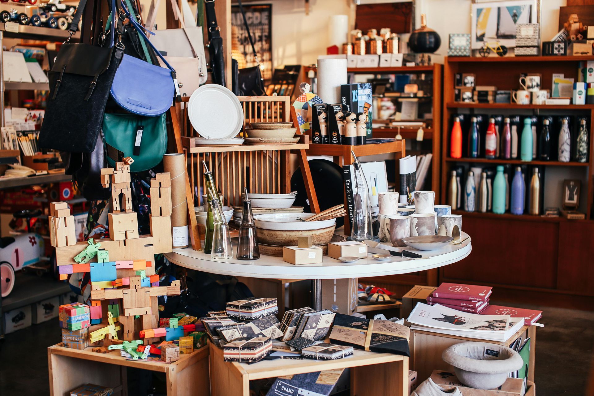 A boutique shop interior with handbags hanging, wooden toys on a crate, a central table of goods, and shelves of bottles.