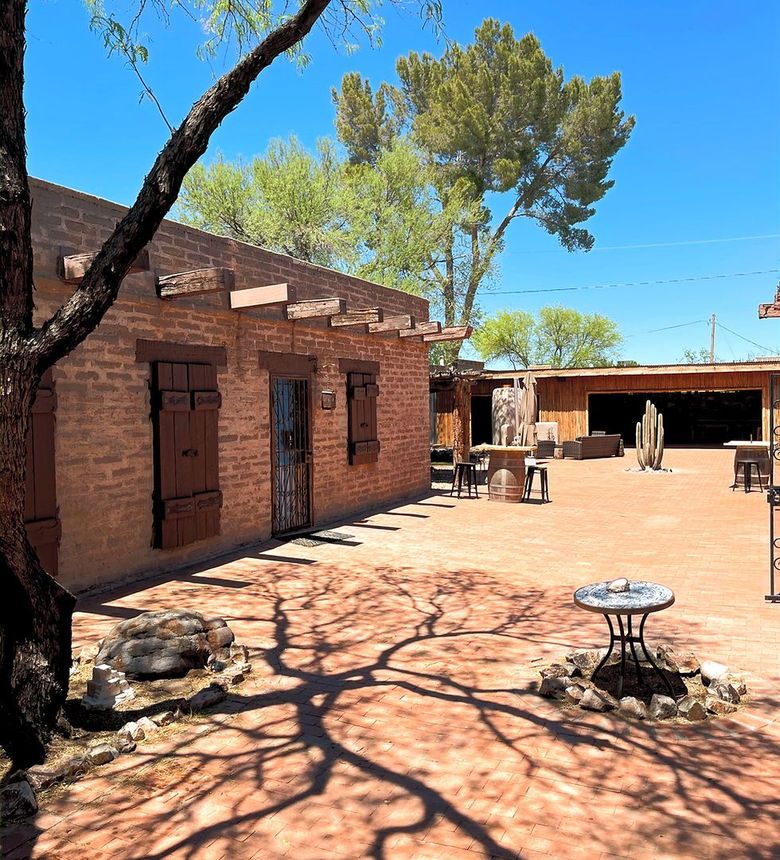 Adobe-style building with courtyard, tree shadows on ground. Bright sunlight, blue sky.
