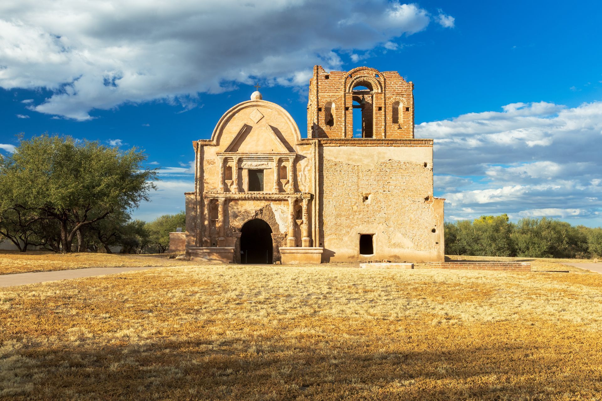 Ruins of a tan adobe church under a bright blue sky with fluffy white clouds and dry, yellow grass.