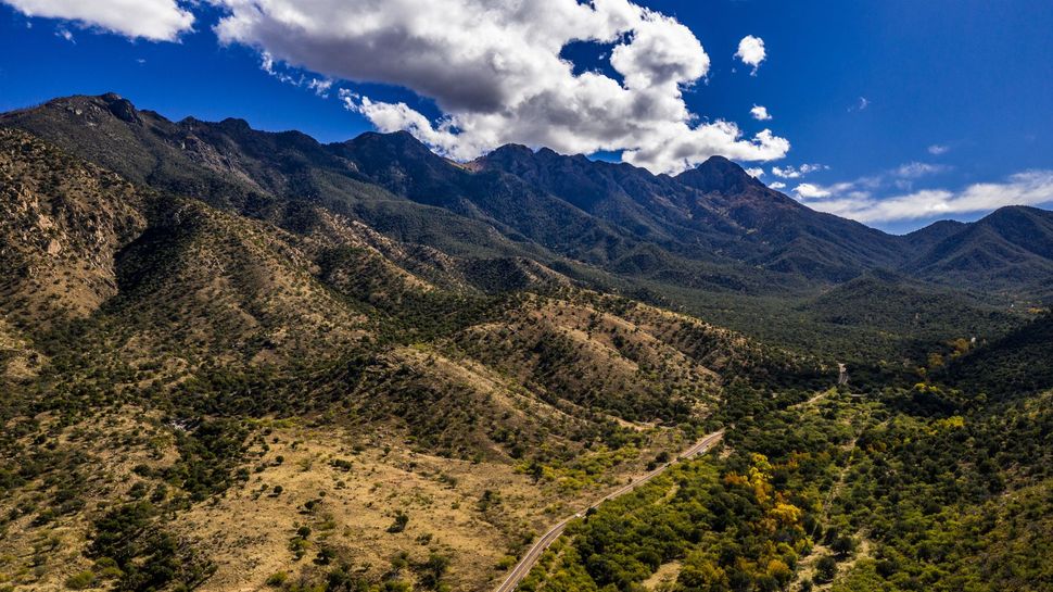 Mountain range under a blue sky with clouds. Green trees cover the slopes, a road winds through the valley.