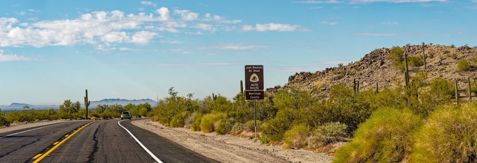 Road through desert landscape with saguaro cacti, sign, and rocky hills under blue sky.