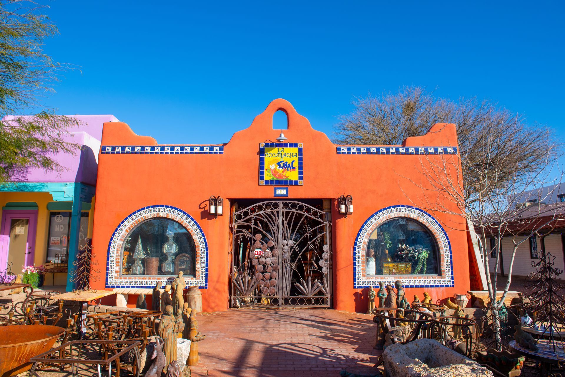 Orange adobe building with decorative iron gate, arched windows, and blue tile trim under a clear sky.