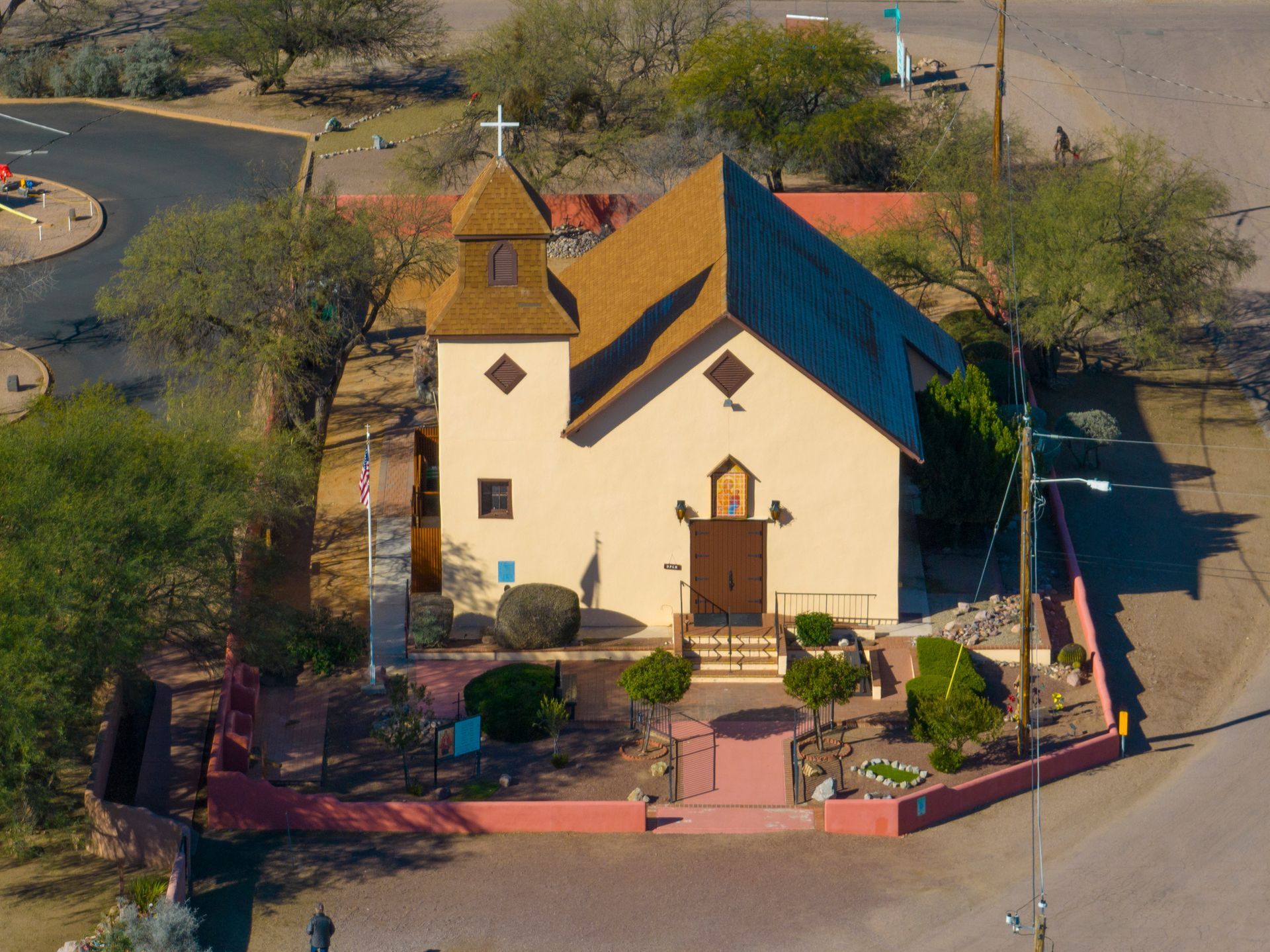 A beige church with brown roof and a cross on top, surrounded by trees and a red brick wall.