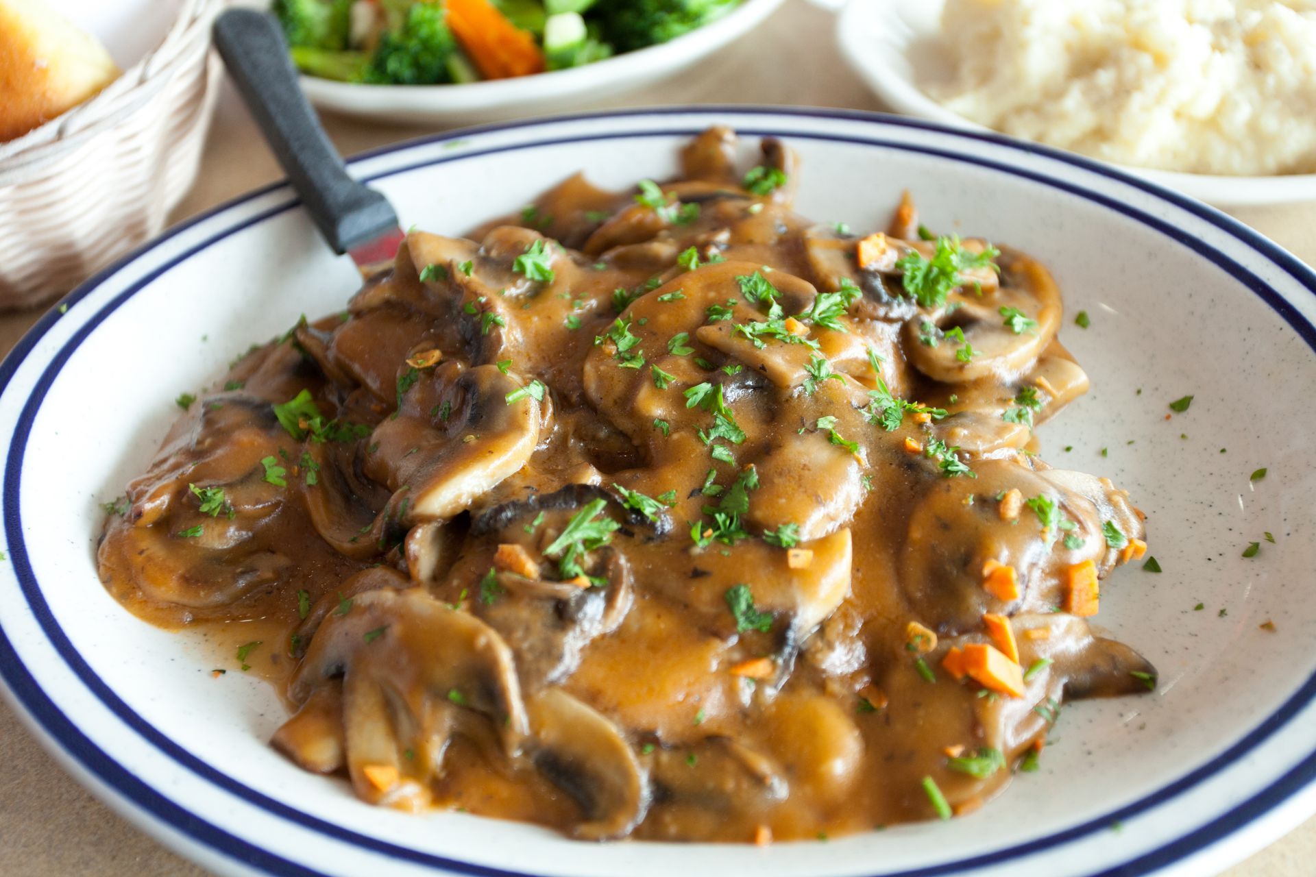 Mushrooms in gravy on a plate, garnished with parsley, with sides of bread, veggies, and mashed potatoes.