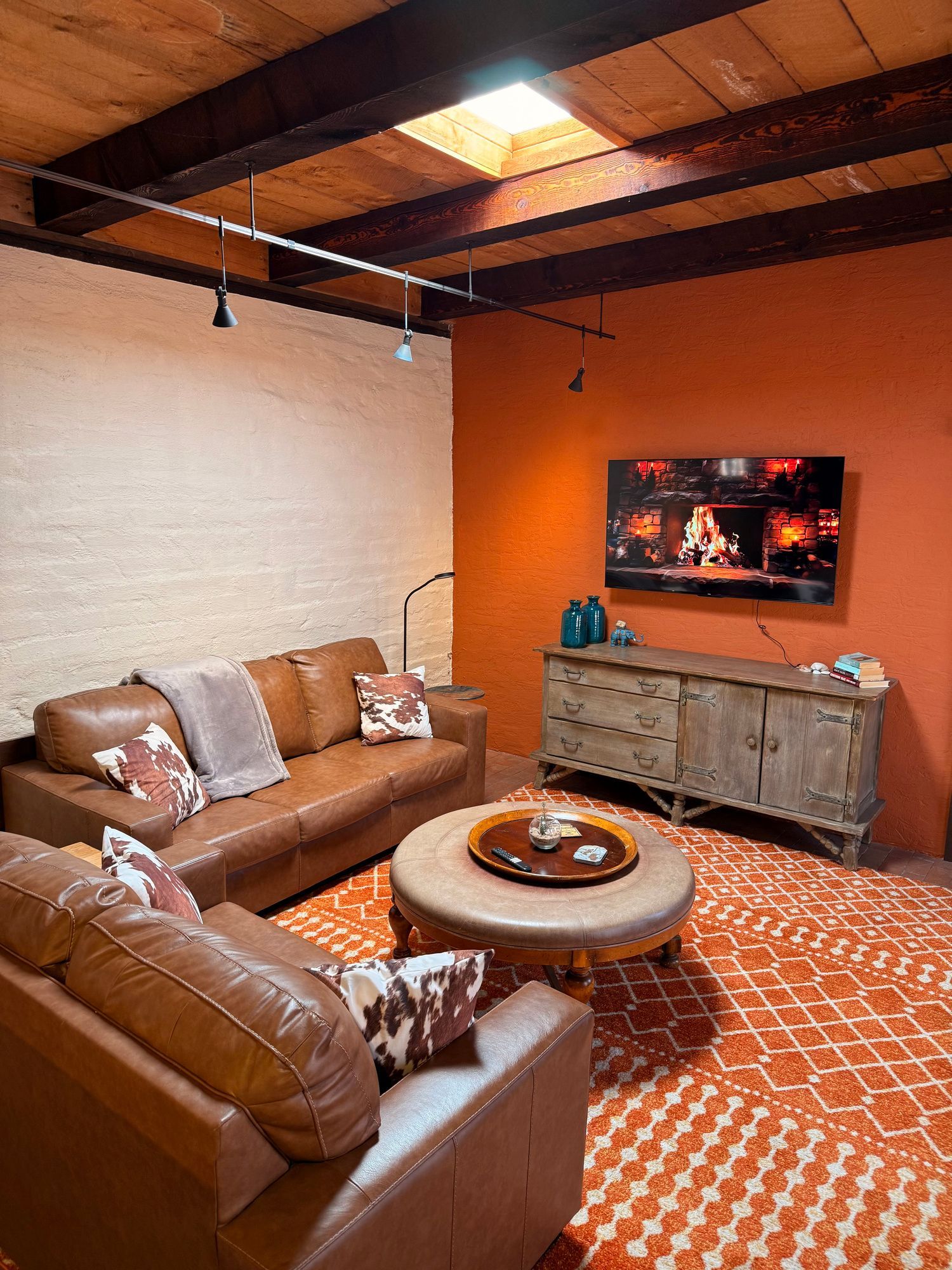 Cozy living room with orange rug, leather sofas, a wooden cabinet, and a TV against a reddish-orange wall.